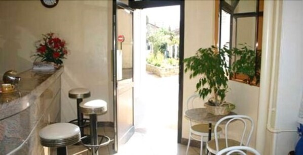 A hotel lobby with a reception desk, two bar stools, a potted plant, and a view of the outside. - Hôtel Le Petit Château (Malakoff)