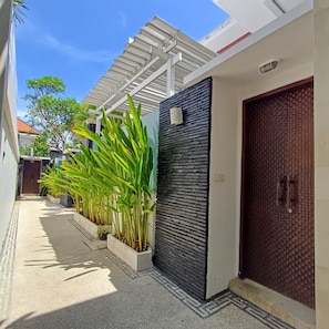 A narrow alleyway with a modern house featuring a dark textured wall, a white door, and a covered walkway.