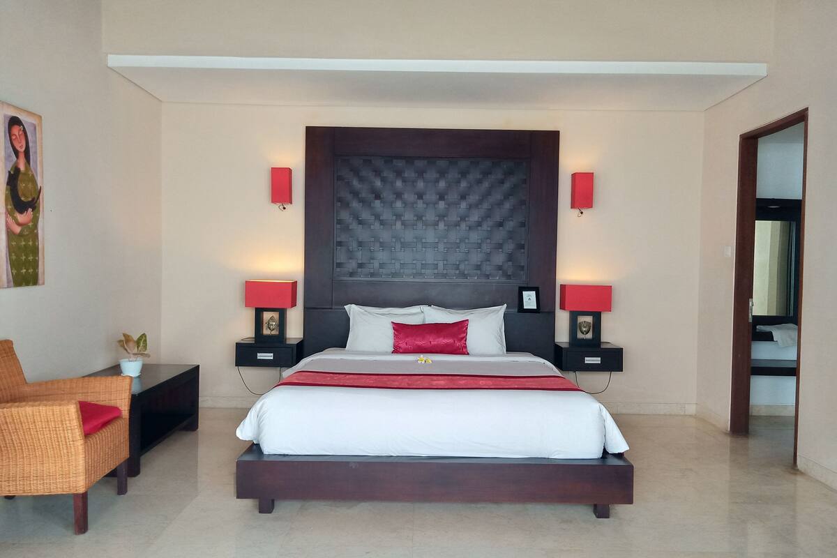 Quarto de hotel moderno com uma cama grande, duas mesas de cabeceira com luminárias, uma mesinha de apoio, uma cadeira de vime e um quadro na parede.