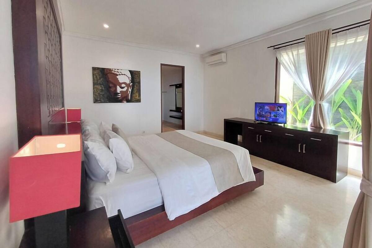 Quarto moderno com uma cama grande, uma TV sobre um móvel de madeira escura e uma janela com cortinas.
