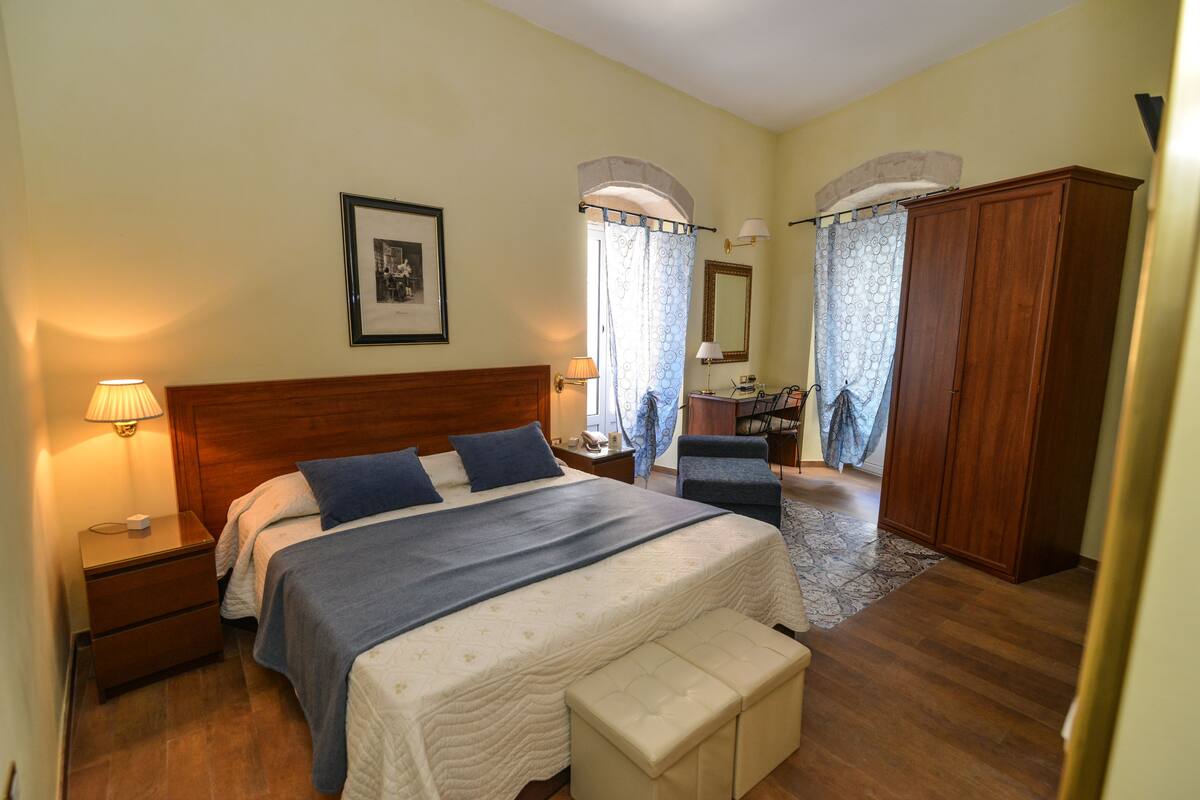 A bedroom with a large bed, a wooden wardrobe, a bedside table with a lamp, and a framed picture on the wall.