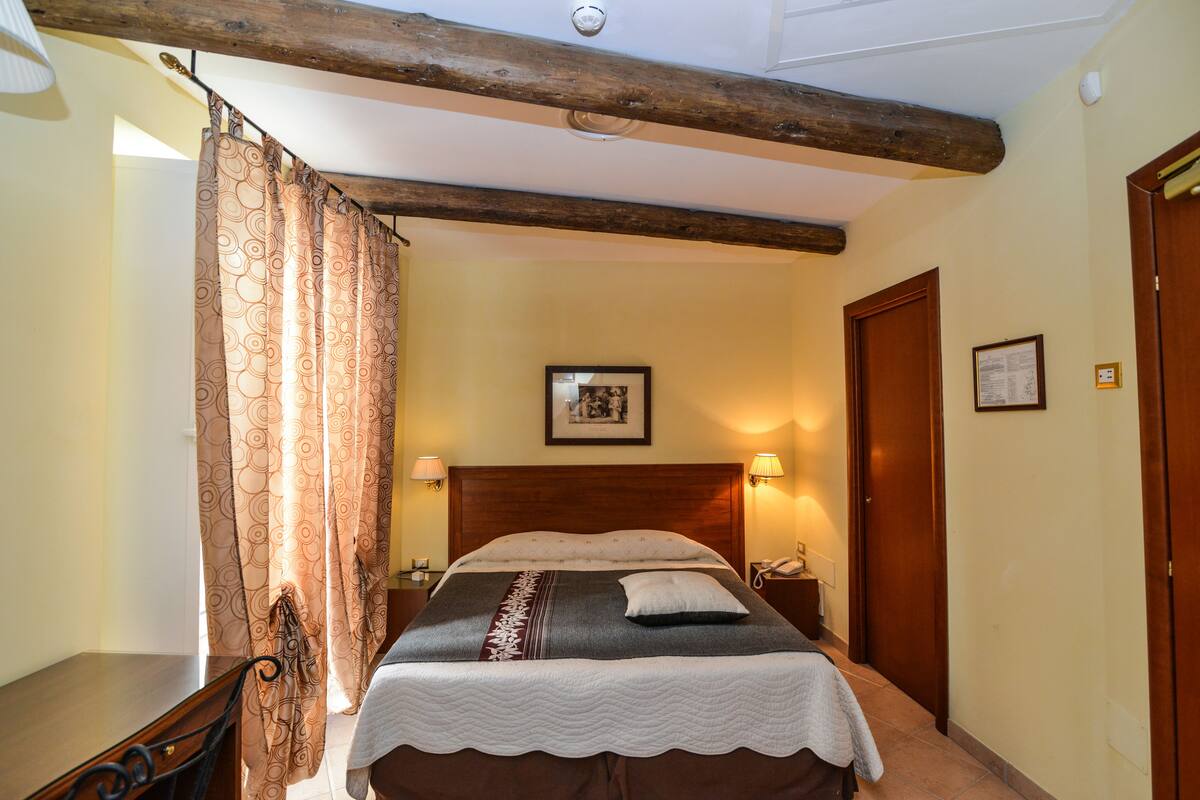 A bedroom with a wooden headboard, a bed with a patterned bedspread, a bedside table with a lamp, and a door to the right.