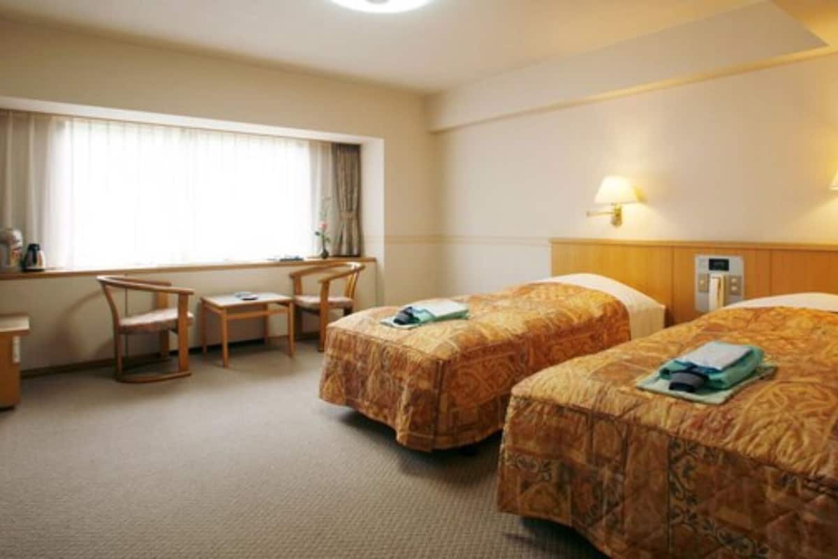 Une chambre d’hôtel avec deux lits, un bureau et une chaise. Elle est équipée d’une fenêtre avec des rideaux, d’une lampe fixée au mur et d’un thermostat.