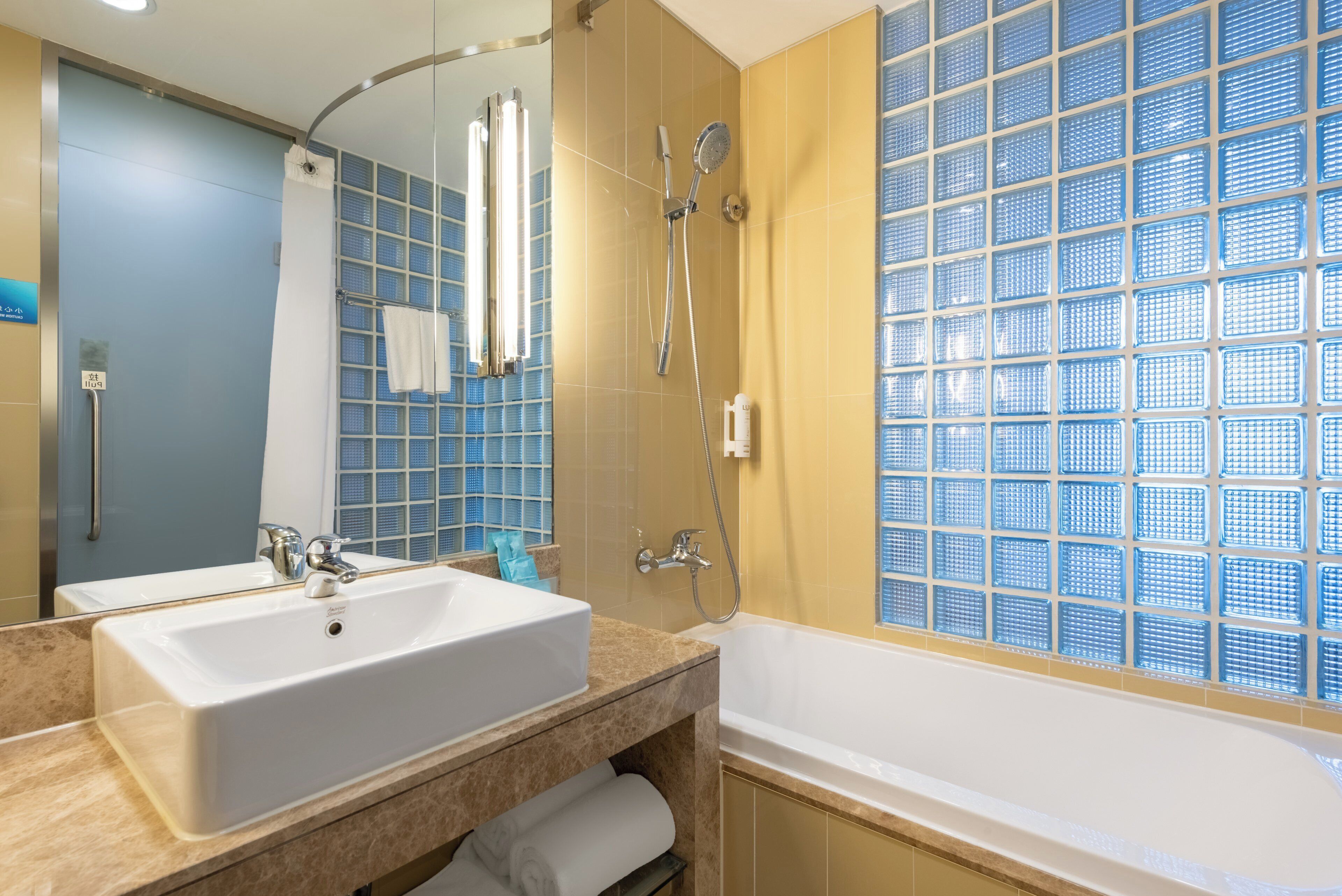 Une salle de bain avec un grand lavabo, un miroir, une douche et une fenêtre ornée d’un motif à carreaux bleus.