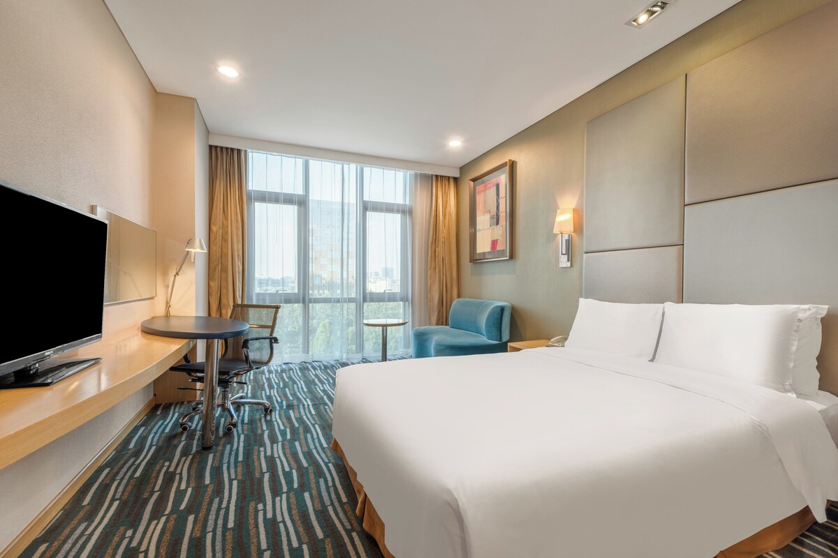 Une chambre d’hôtel comprenant un lit, un bureau avec une télévision, une chaise, une petite table et une fenêtre avec des rideaux.