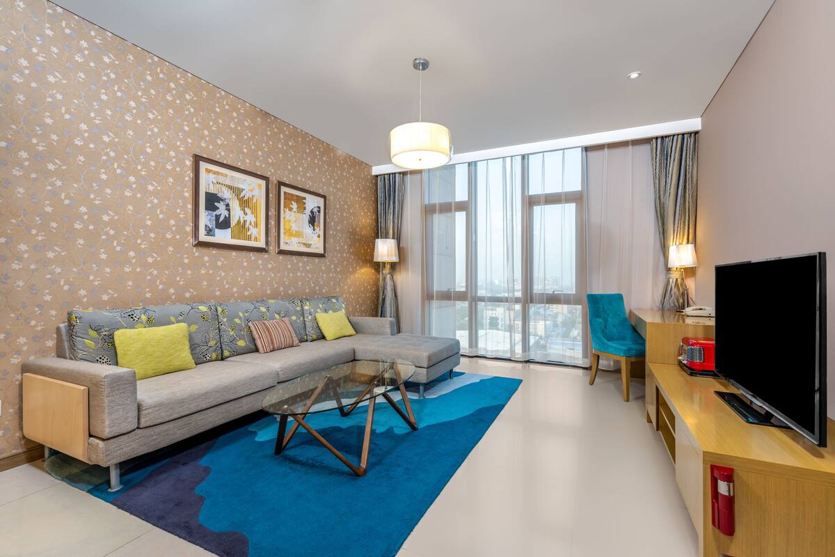 A modern hotel room with a sofa, a coffee table, a TV, and a large window.