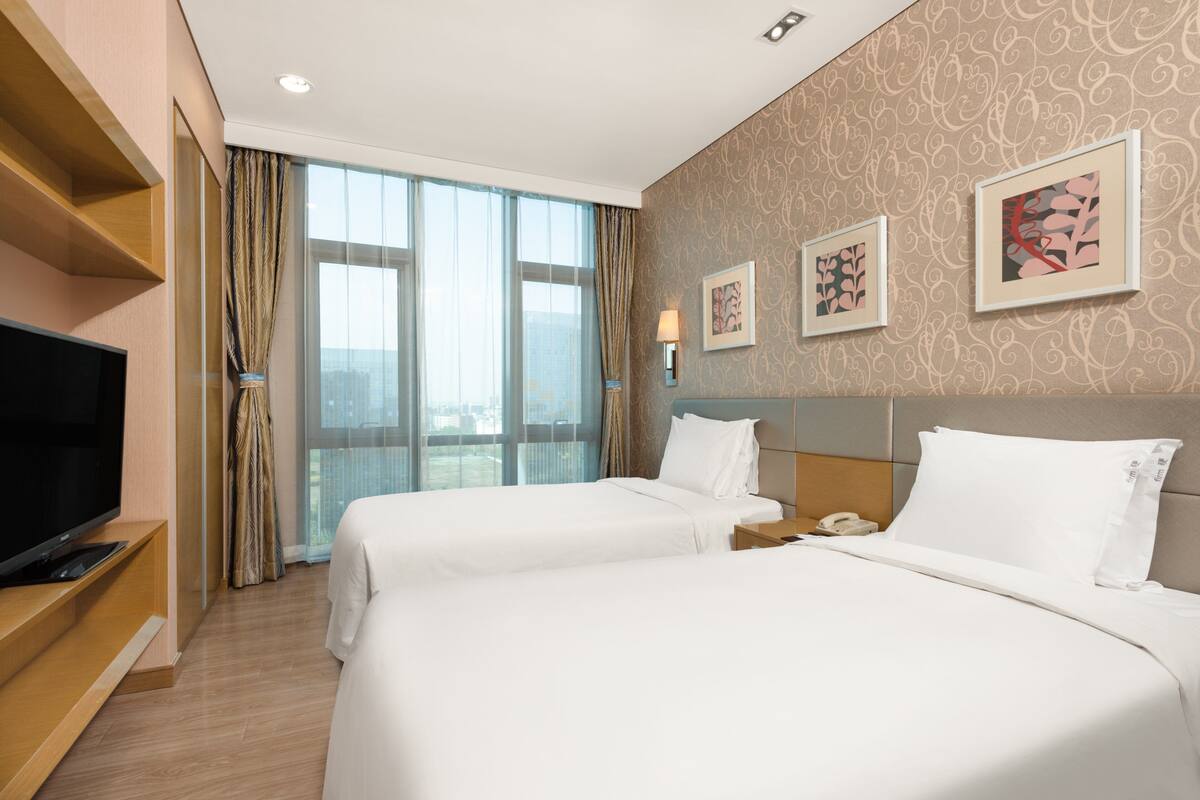 A hotel room with two beds, a TV, a desk, and a window with curtains.