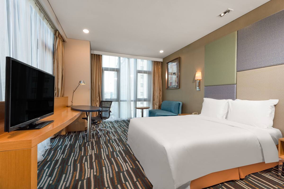 A hotel room with a large bed, a desk with a flat-screen TV, a small table, and a chair.