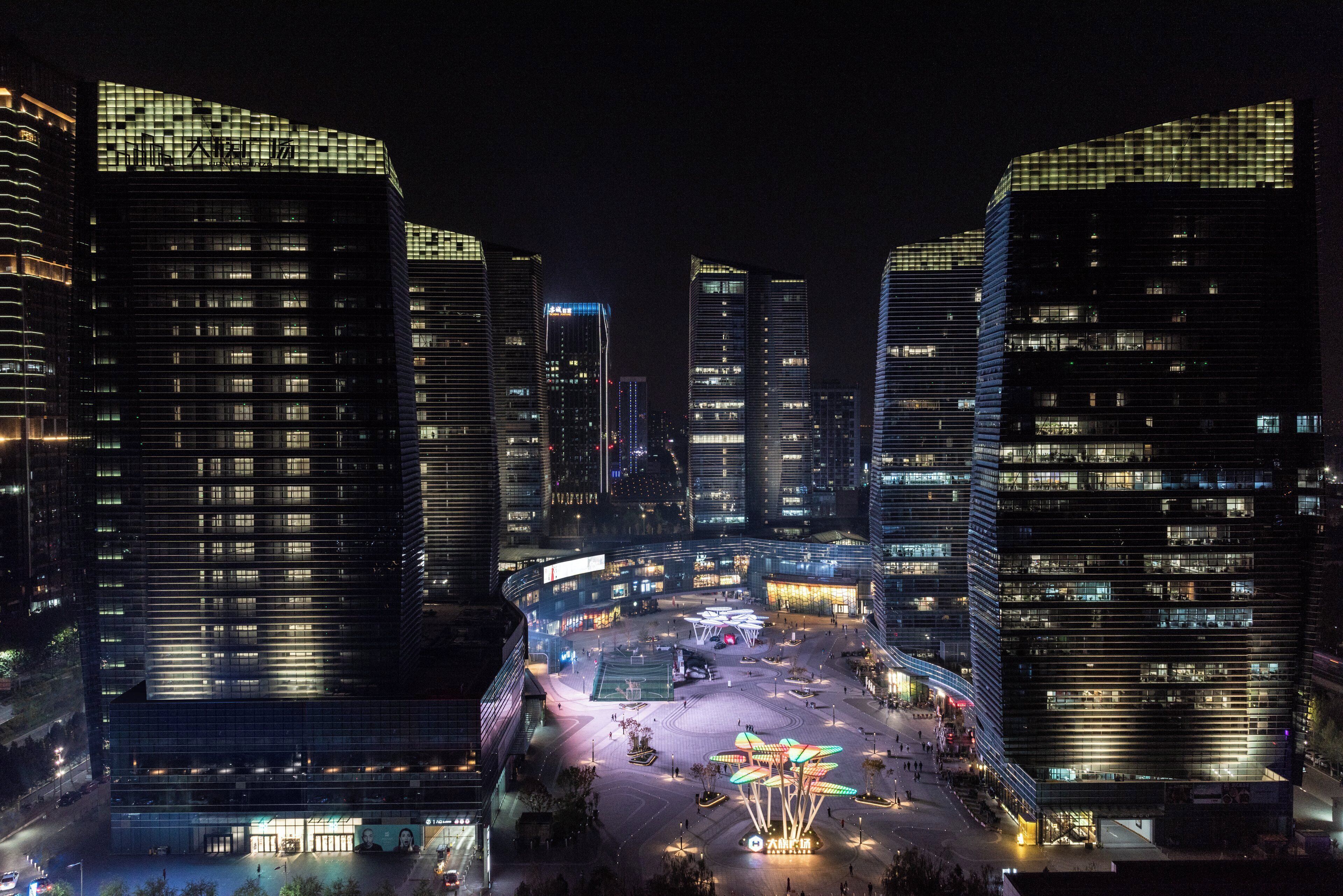 A nighttime cityscape with modern high-rise buildings and a central plaza with a unique light installation.