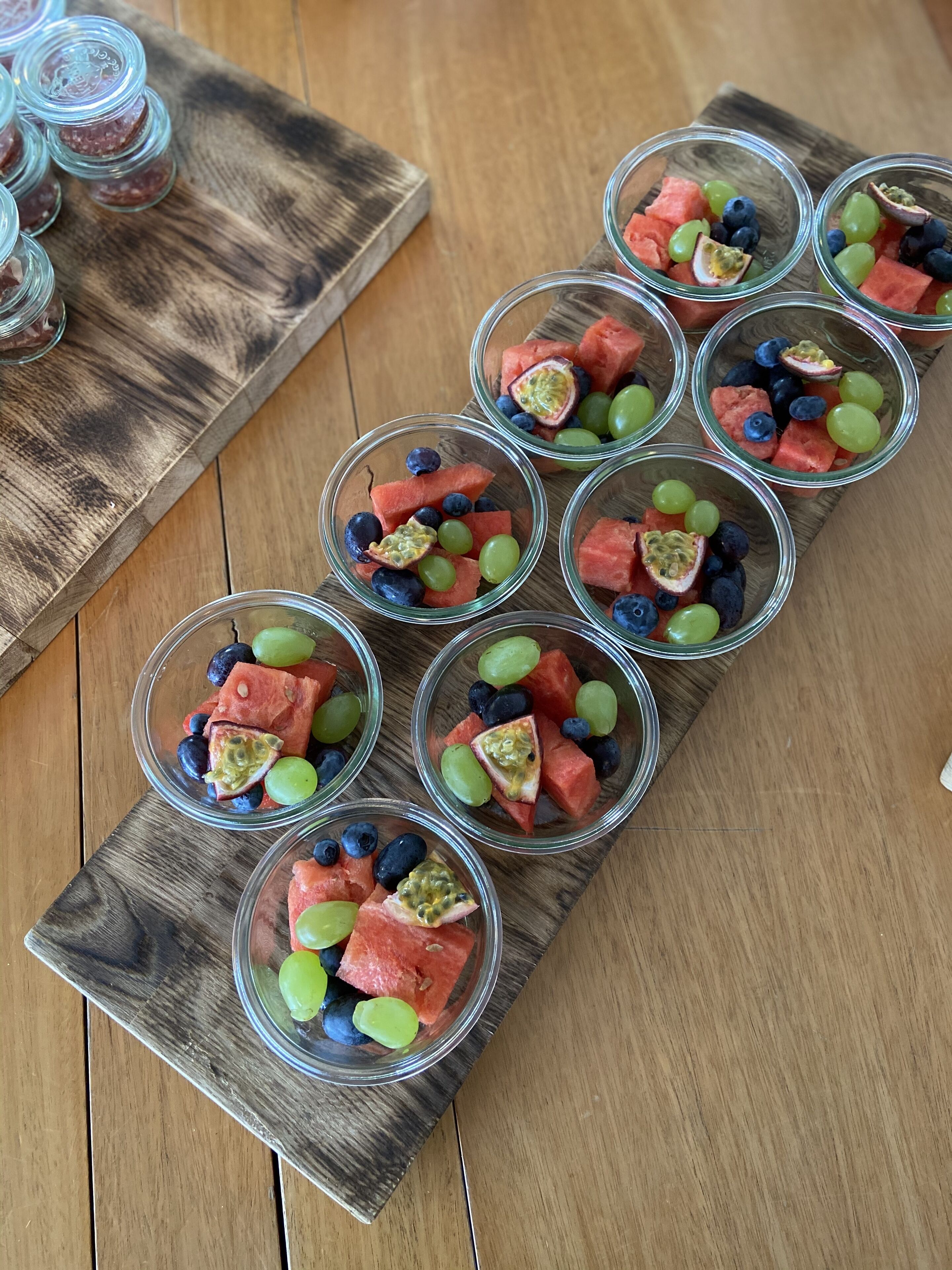 A wooden serving board with clear glass cups containing watermelon, grapes, and passion fruit.