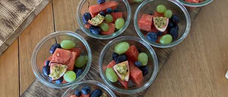 A wooden serving board with clear glass cups containing watermelon, grapes, and passion fruit.