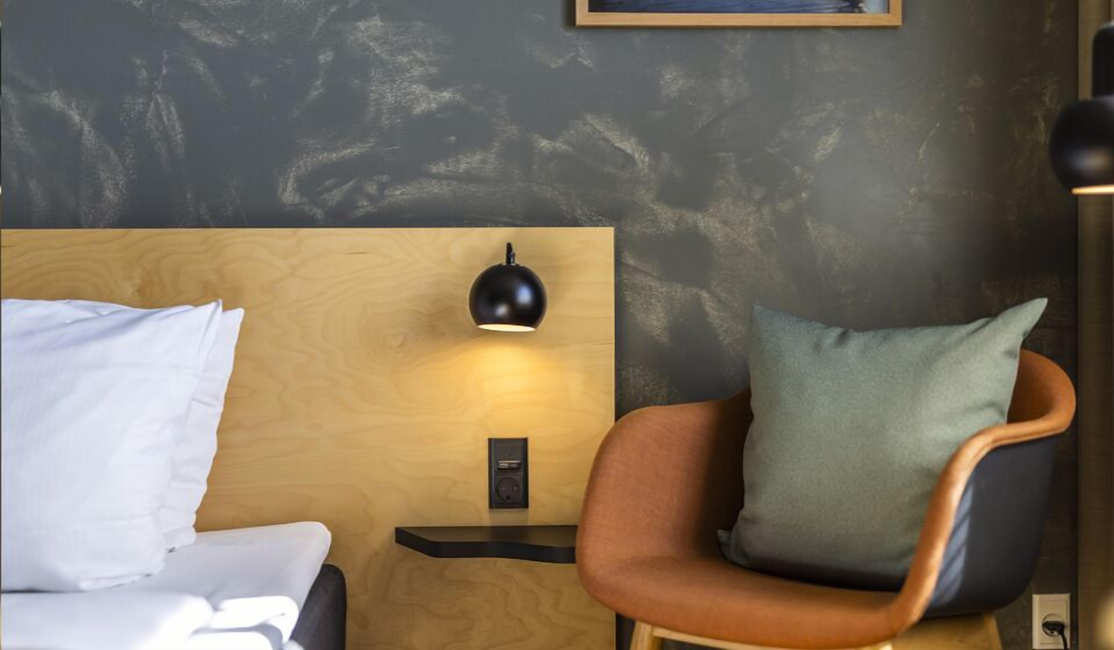 Una habitación de hotel con cabecera de madera, una lámpara negra de pared, una mesita de noche pequeña y una silla con almohada verde.