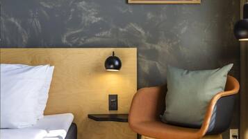 Una habitación de hotel con cabecera de madera, una lámpara negra de pared, una mesita de noche pequeña y una silla con almohada verde.