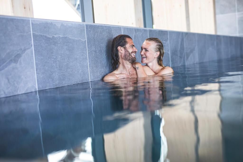 Un couple se trouve dans une piscine intérieure moderne, avec un mur carrelé et un fond en verre transparent.