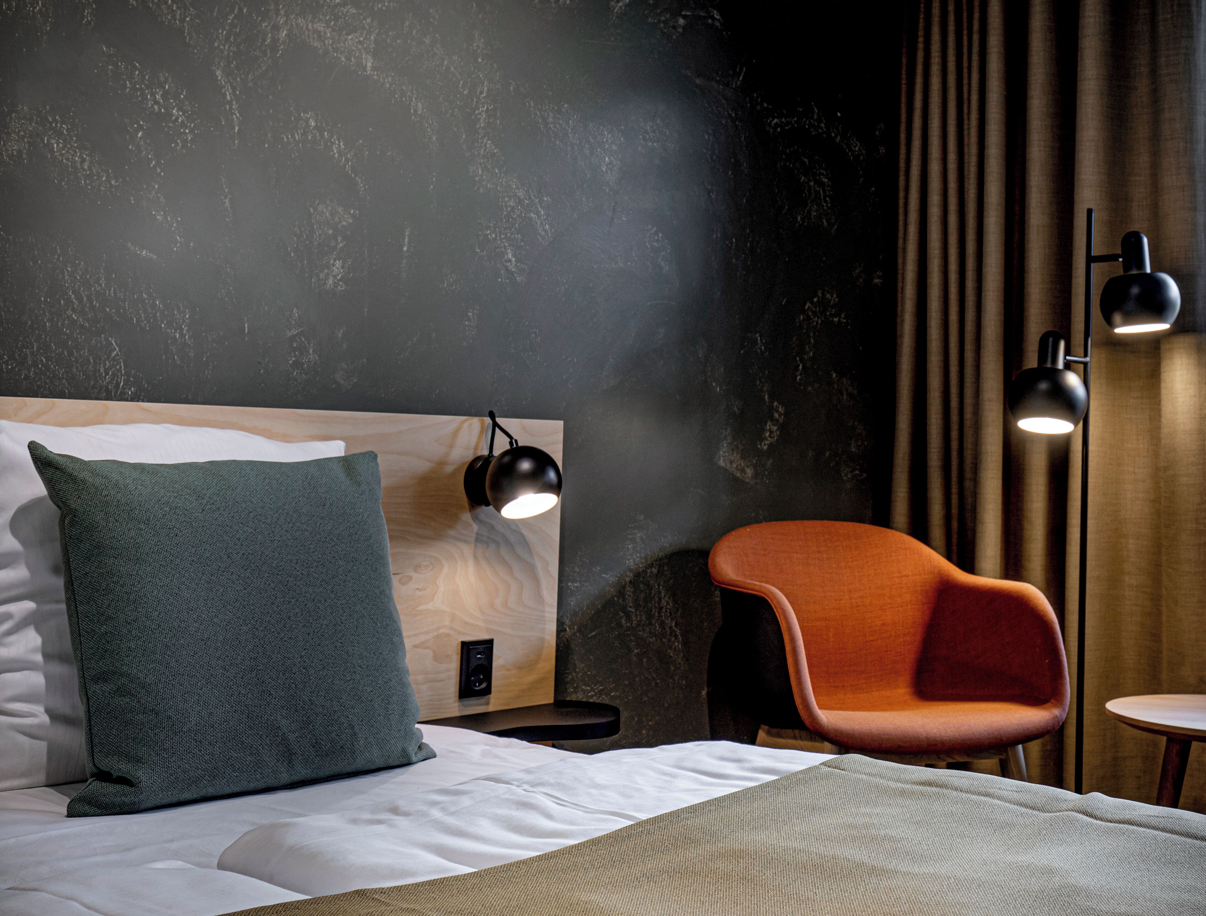 A modern hotel room with a wooden headboard, a green pillow, a wooden chair, and two black floor lamps.