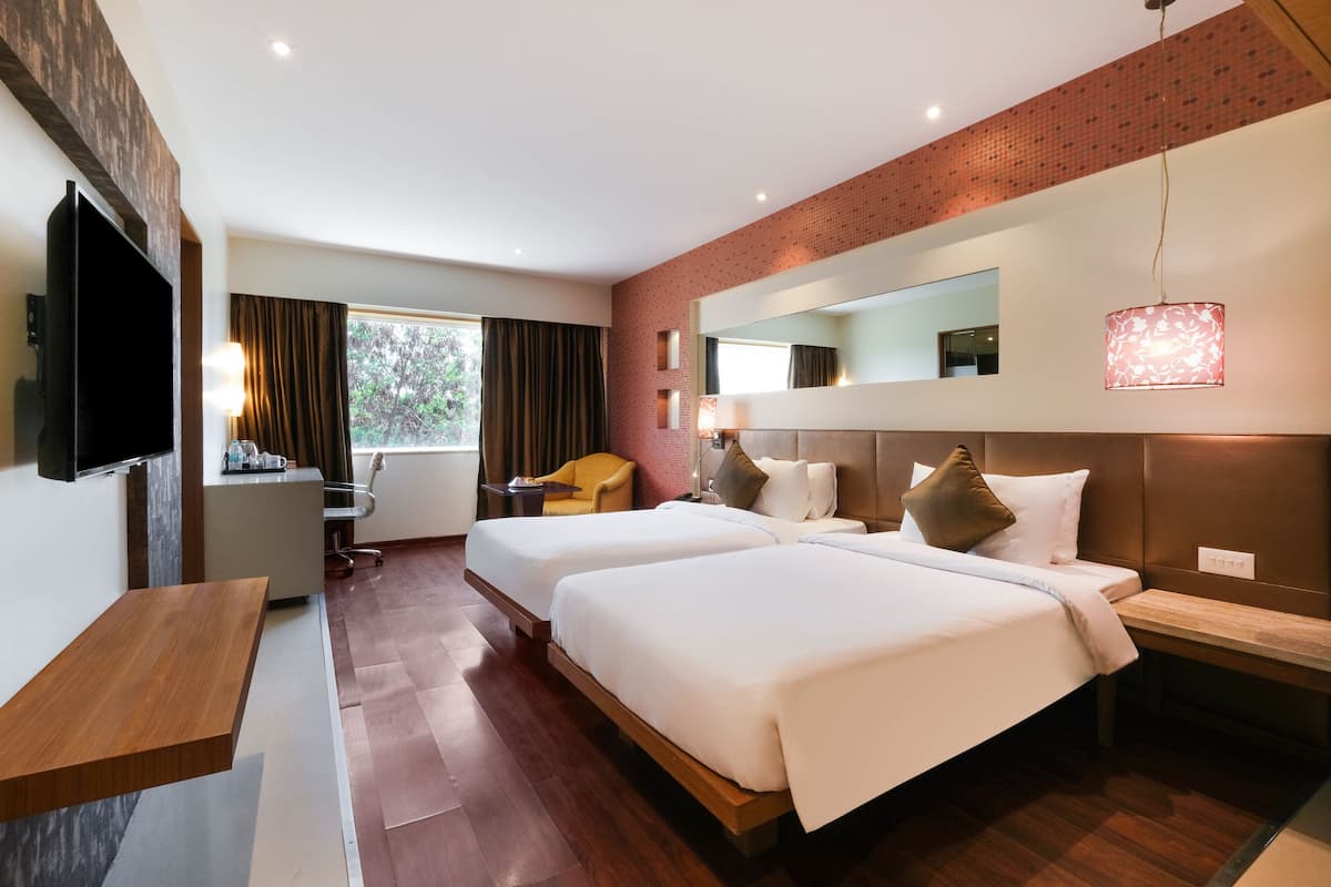 A hotel room with two beds, a TV, a desk, a chair, a window with curtains, and a brick wall.