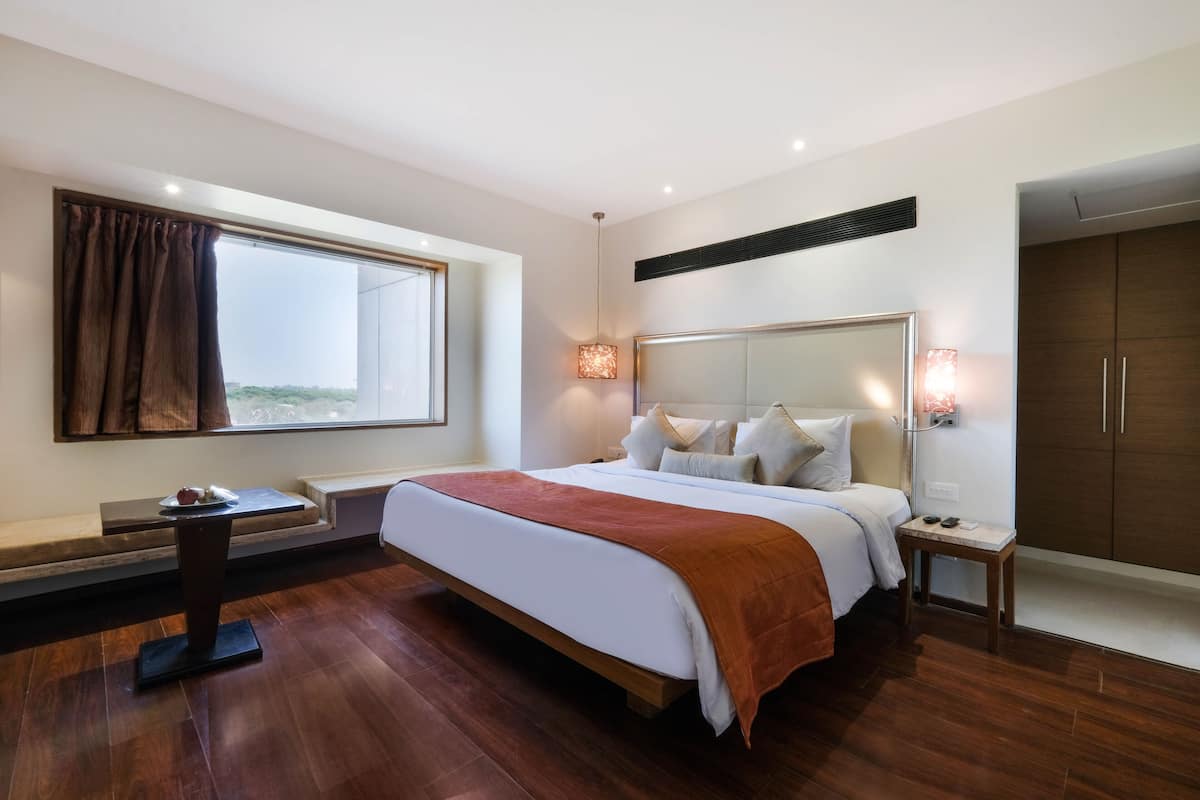 A modern hotel room with a large bed, a bedside table, a nightstand, a window with curtains, a wardrobe, and a built-in heater.