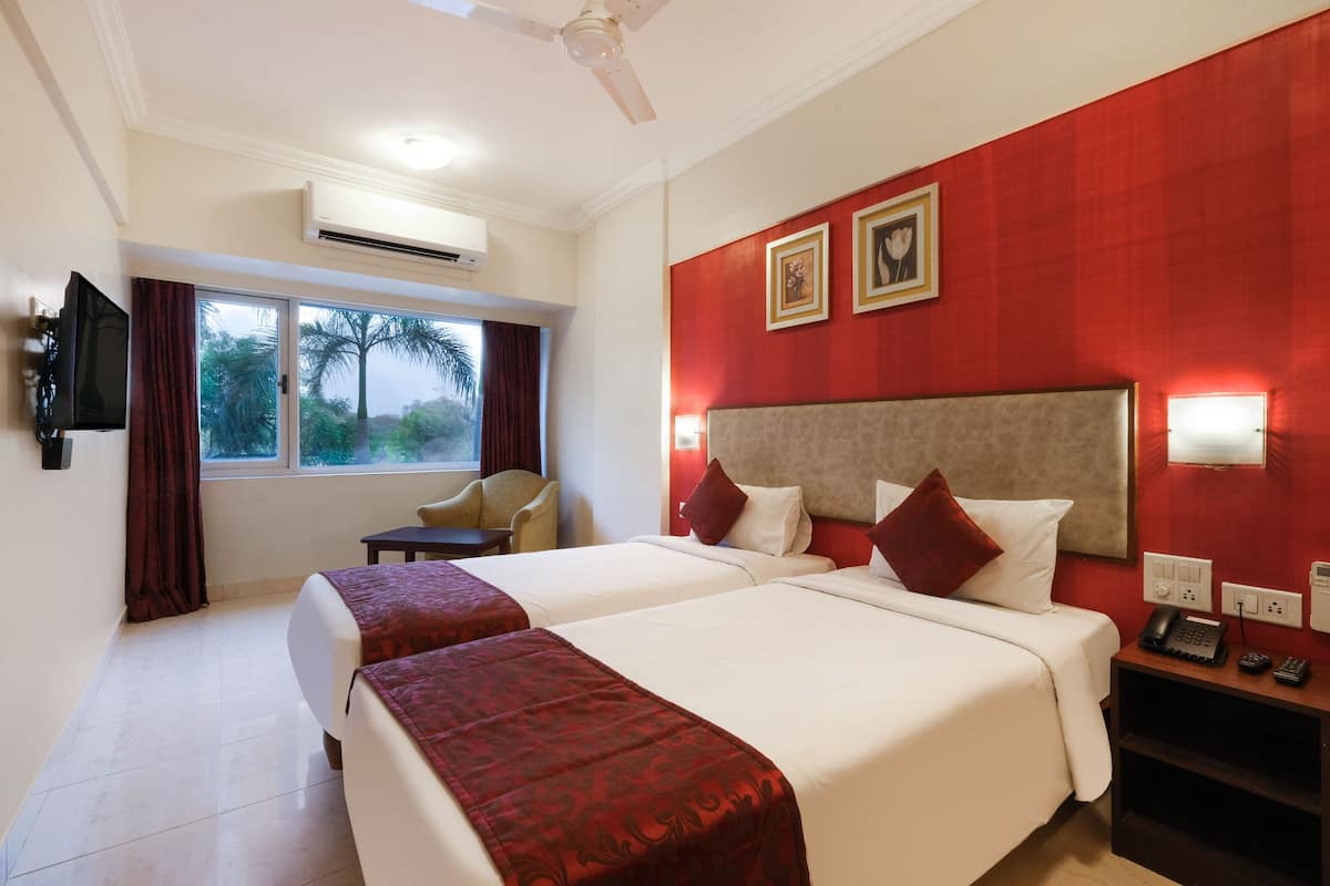 A hotel room with two beds, a TV, a desk with a phone, and a window with a view of trees.
