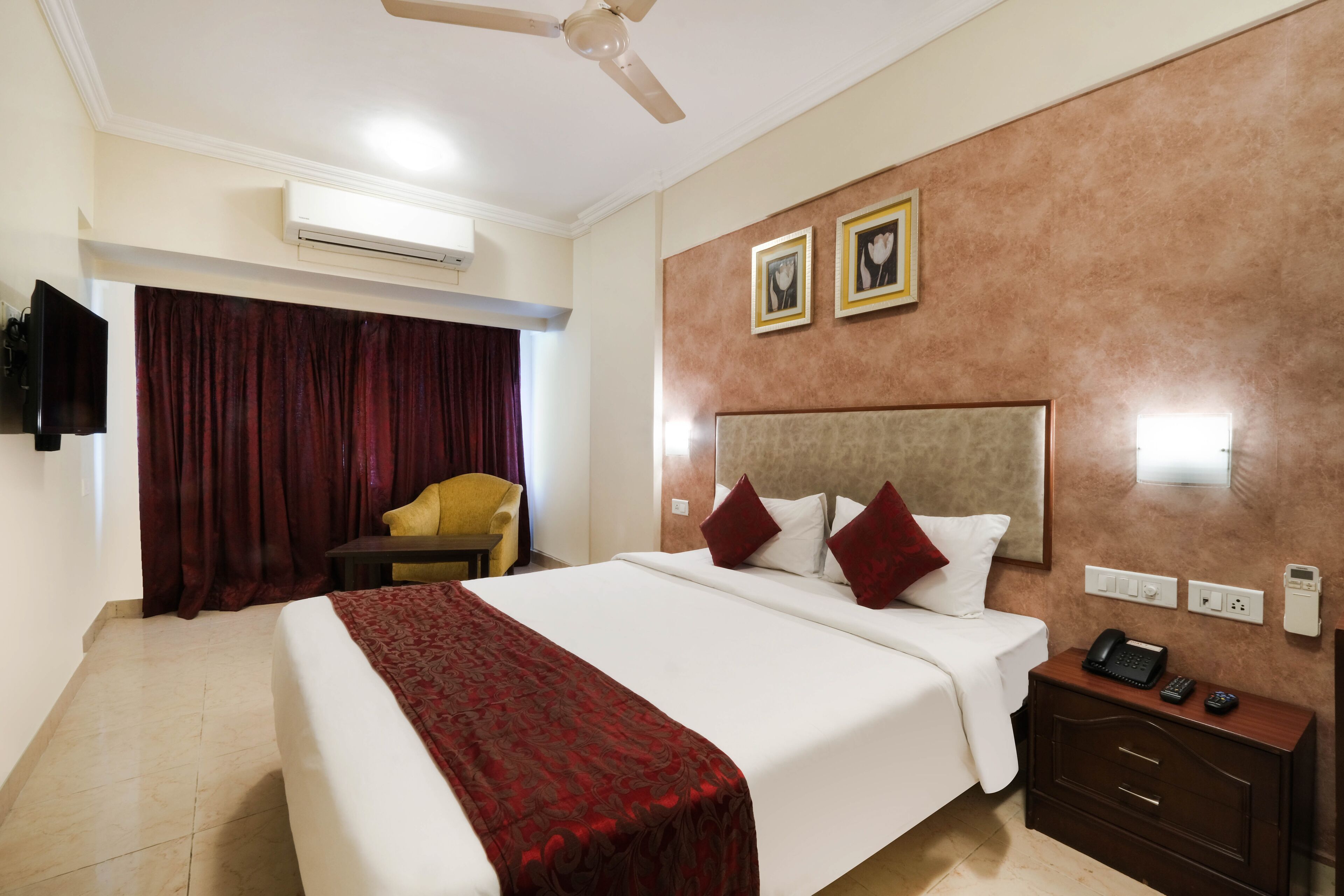 Quarto de hotel com uma cama grande, uma mesinha de cabeceira com telefone e controles remotos, um aparelho de ar condicionado, um ventilador de teto, uma TV fixada na parede e uma cortina vermelha.