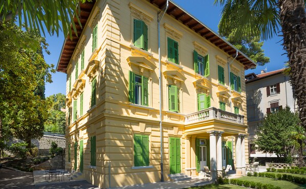 Un edificio amarillo con contraventanas verdes y un balcón, rodeado de vegetación.