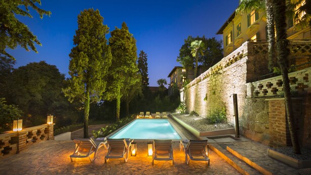 Uma piscina com espreguiçadeiras e lanternas, cercada por árvores e um muro de pedra.