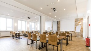 A spacious conference room with multiple tables and chairs, large windows, and a stone accent wall. - RiKu HOTEL Neu-Ulm (Neu-Ulm)