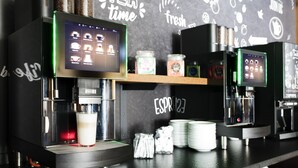 A coffee shop counter with espresso machines, a digital menu display, and a chalkboard wall with various coffee-related drawings. - RiKu HOTEL Neu-Ulm (Neu-Ulm)