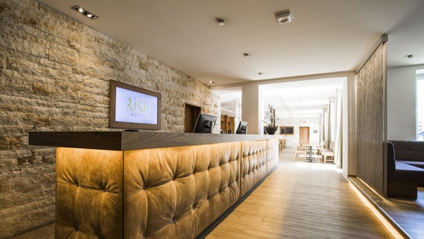 A reception area with a stone wall, a wooden reception desk, and a view into the hotel lobby. - RiKu HOTEL Neu-Ulm (Neu-Ulm)