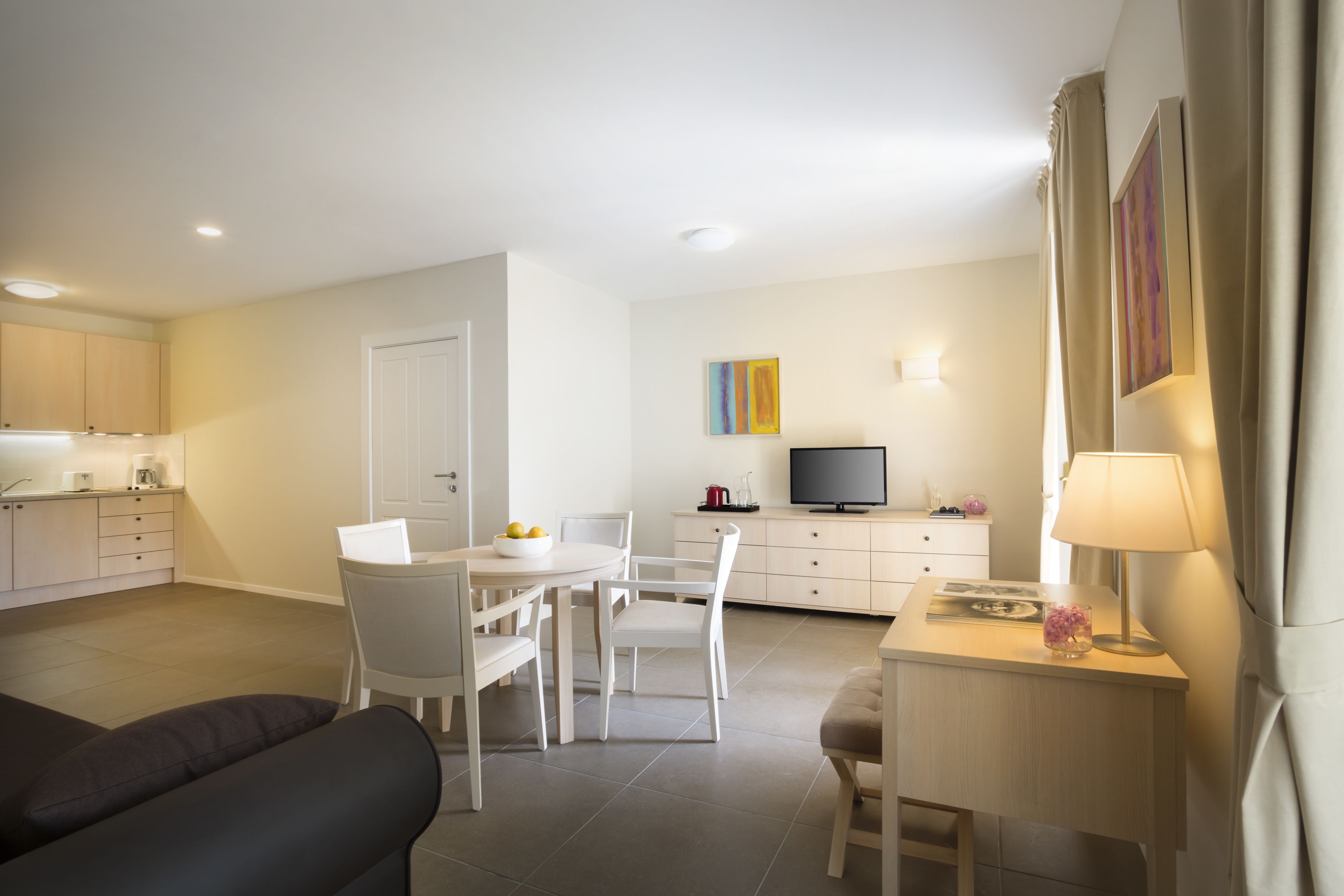 Una sala de estar moderna con un sofá, una mesa de comedor con sillas, un televisor y un escritorio pequeño con una lámpara.