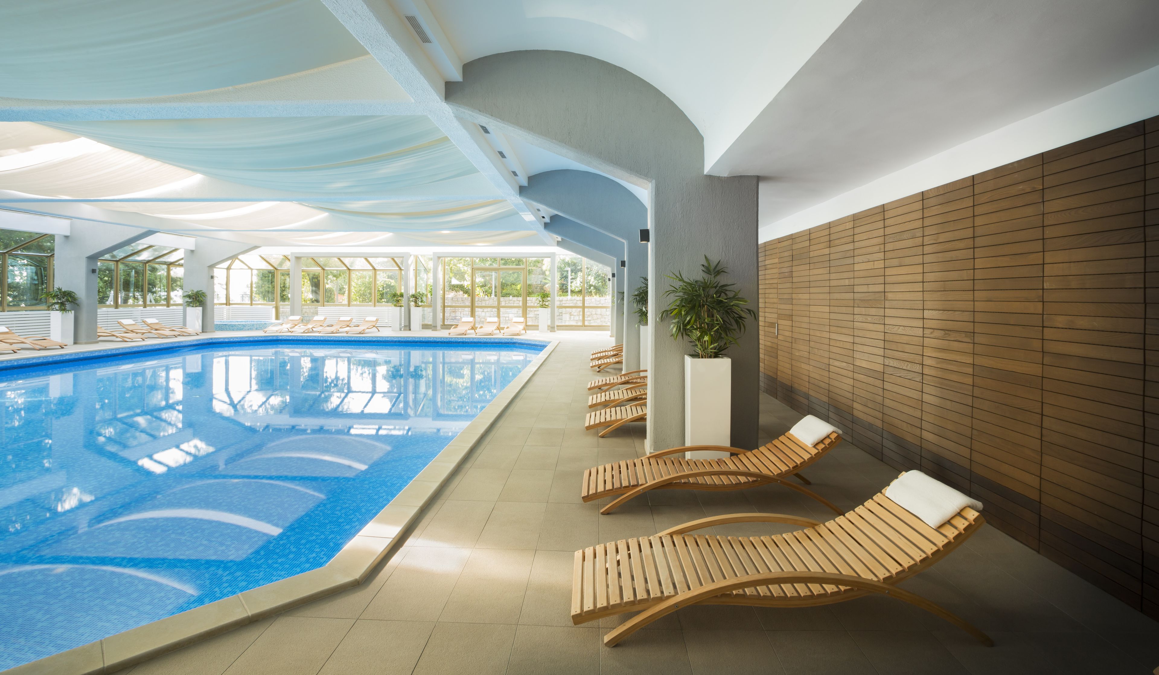 A modern indoor swimming pool with clear blue water, wooden loungers, and a covered area with natural light.