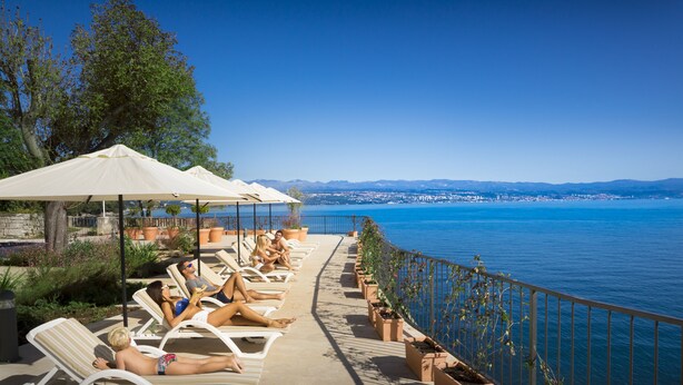 A seaside terrace with sun loungers and umbrellas overlooking the sea and a distant cityscape. - Apartments Belvedere - Liburnia (Lovran)