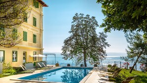 A pool area with lounge chairs, a large tree, and a view of the sea. - Apartments Belvedere - Liburnia (Lovran)