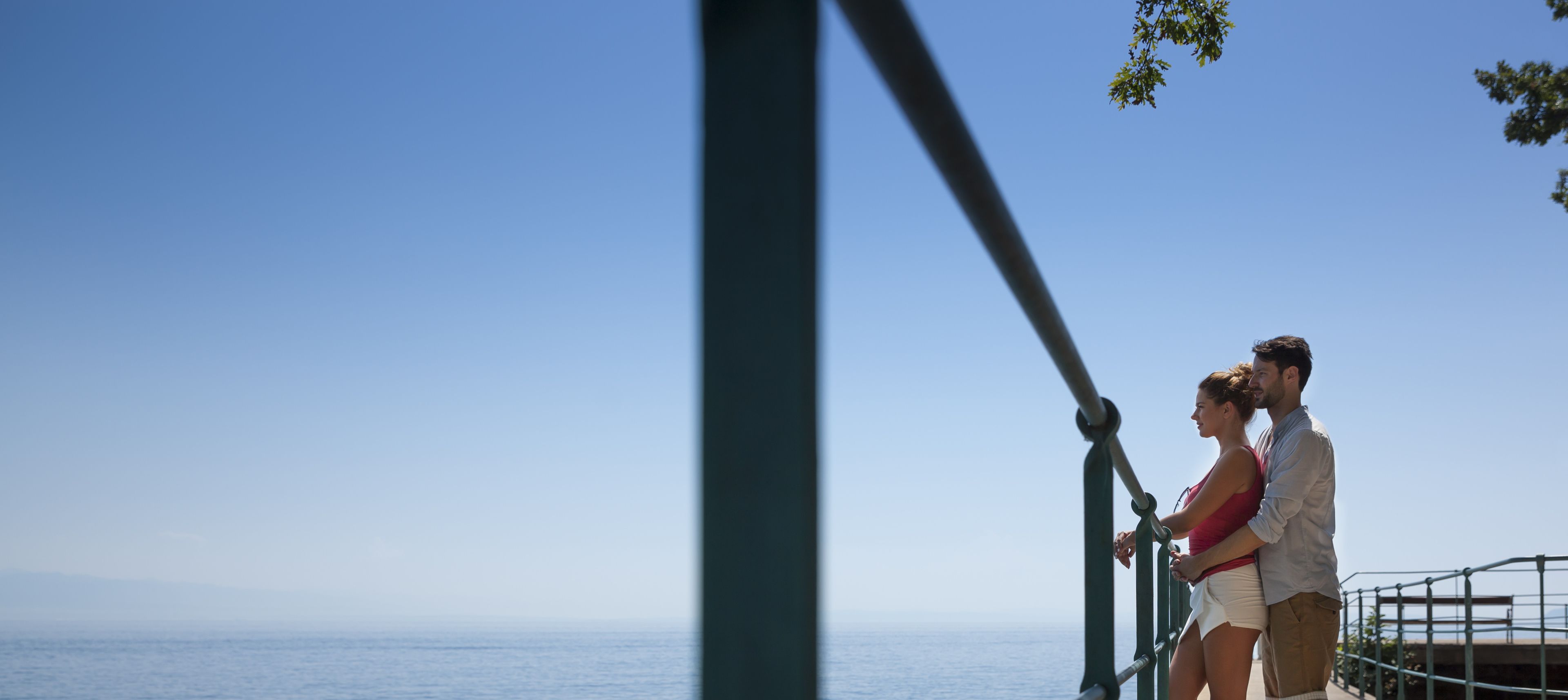 Una pareja está de pie junto a una barandilla con vistas a un cuerpo de agua, con un cielo despejado al fondo.