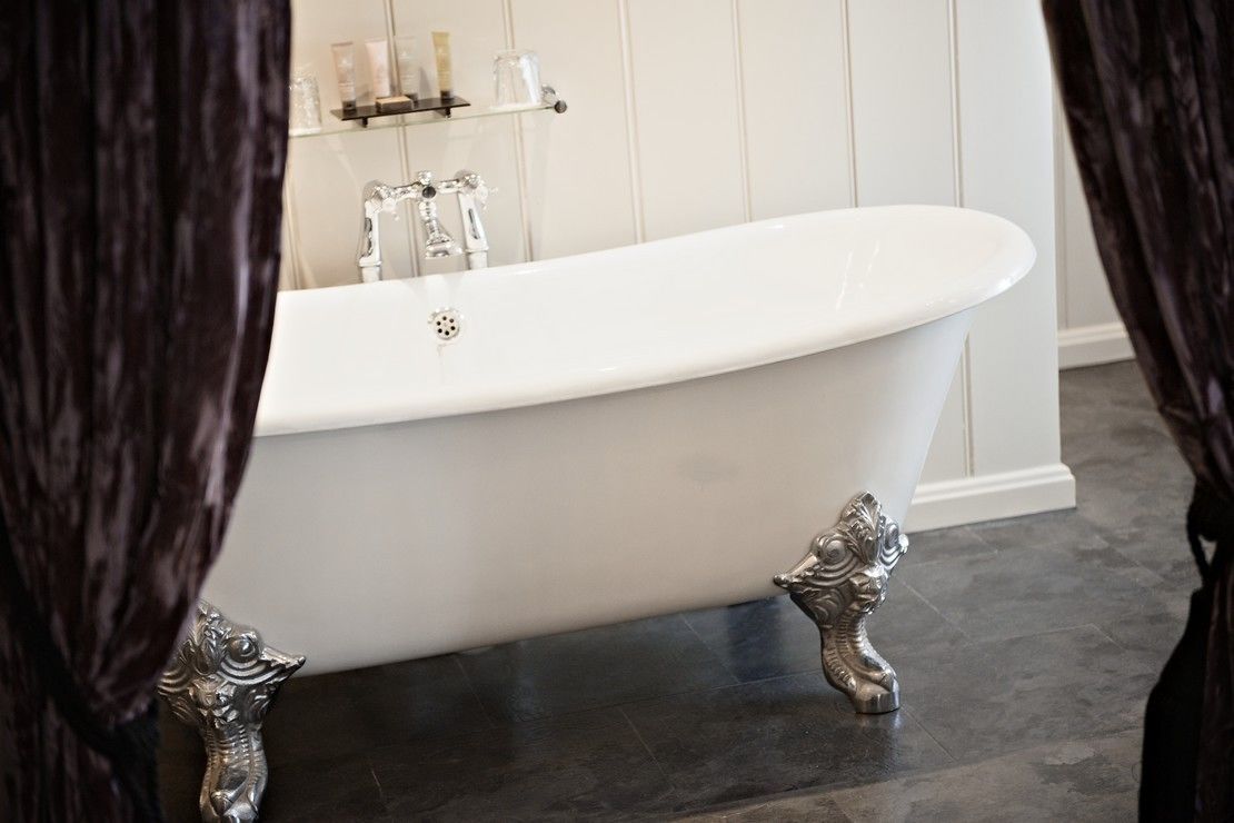 Een wit, vrijstaand bad met decoratieve pootjes, in een badkamer met donkere betegelde vloer en witte wanden.
