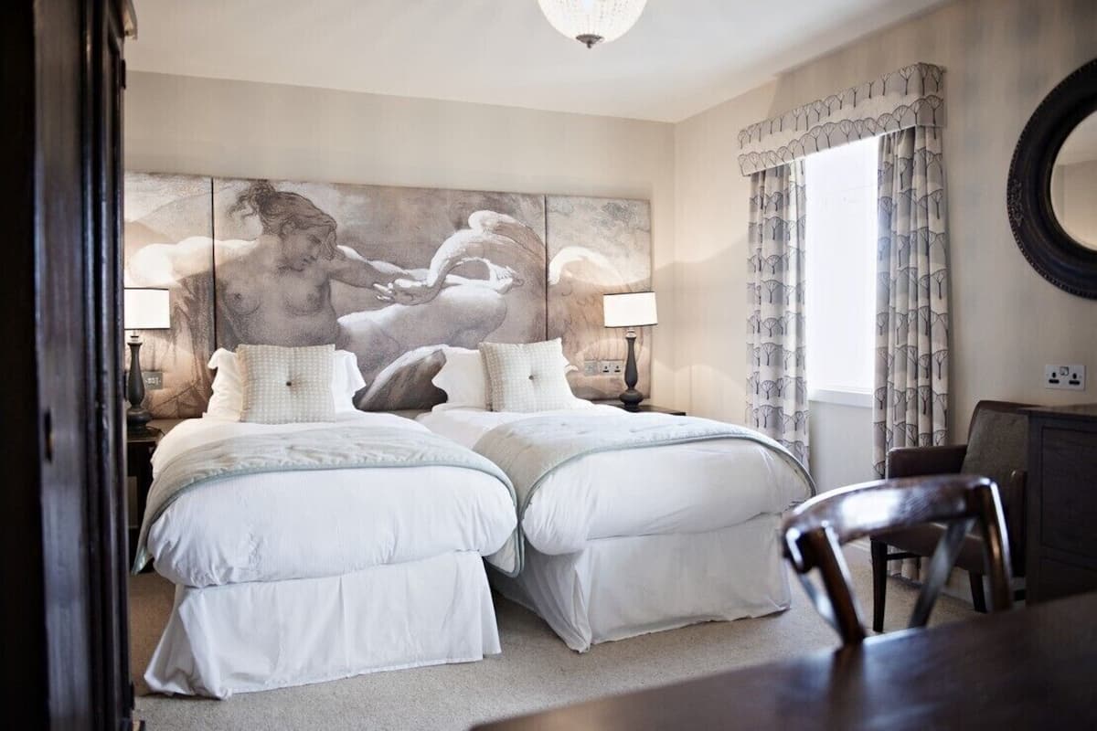 Une chambre avec deux lits, un grand tableau au mur, un miroir rond et une fenêtre avec des rideaux.