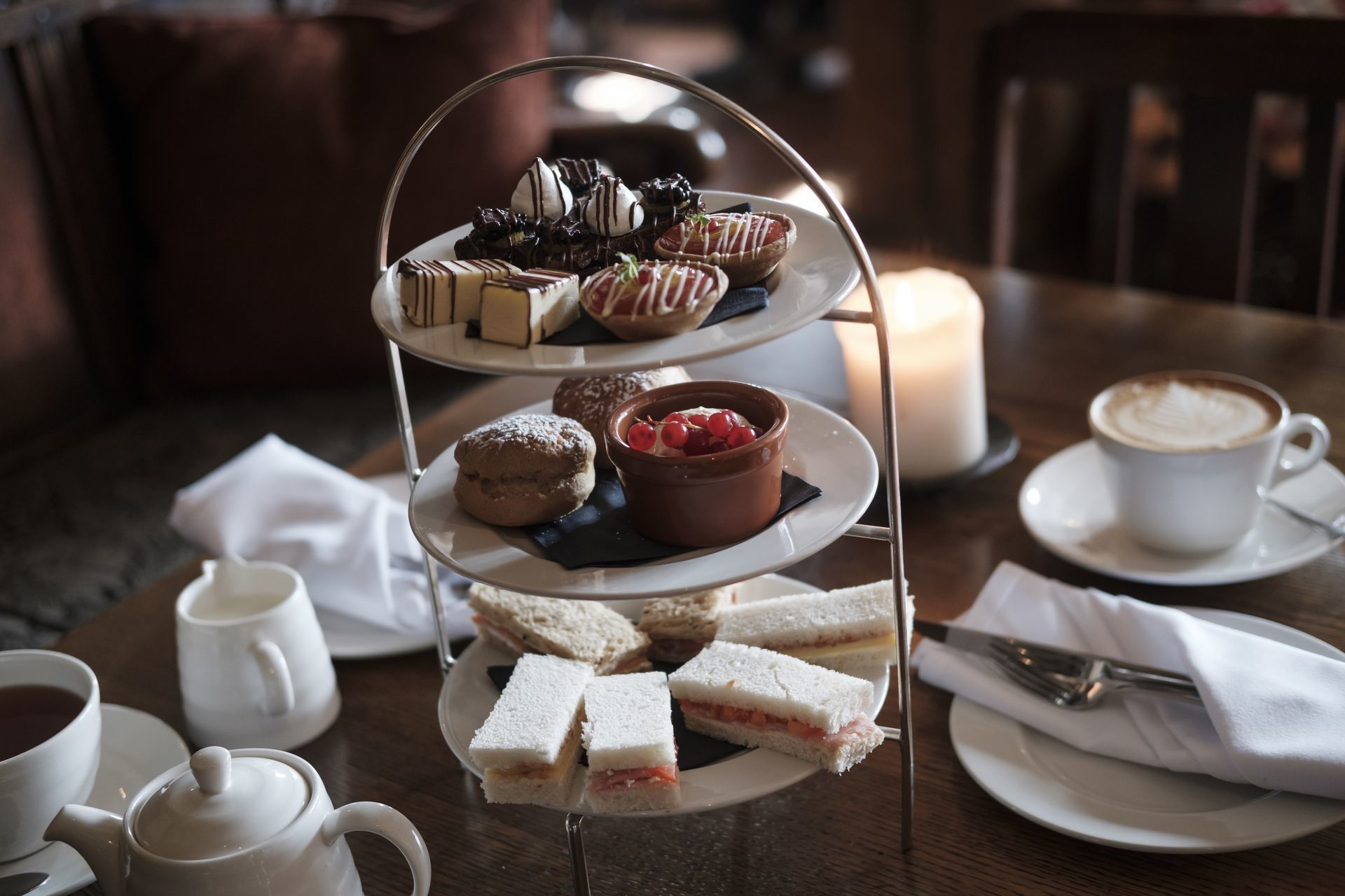 A three-tiered dessert stand with assorted pastries and sandwiches, a cup of coffee, and a teapot on a wooden table.