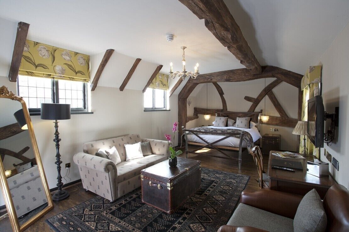 A rustic bedroom with exposed wooden beams, a large bed, a sofa, a chest of drawers, and a mirror.