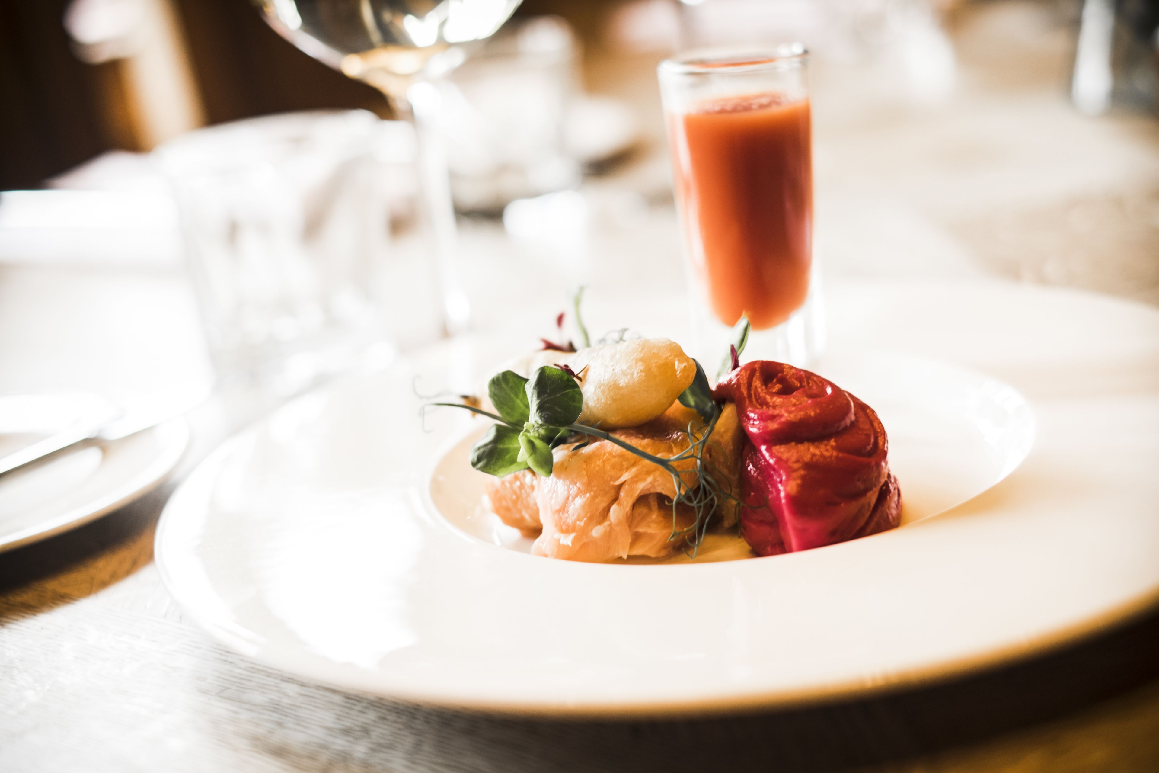 A white plate with a dessert consisting of a red rose, a yellow item, and some greens, accompanied by a glass of red liquid.