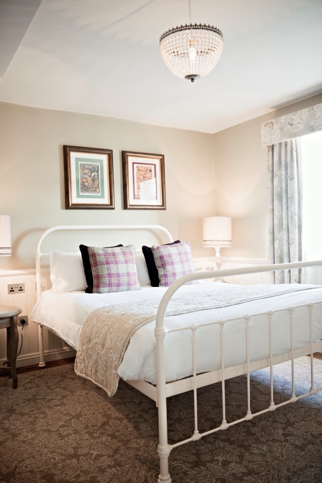 A bedroom with a white metal bed frame, a chandelier, two framed pictures on the wall, a bedside table with a lamp, and a window with curtains.