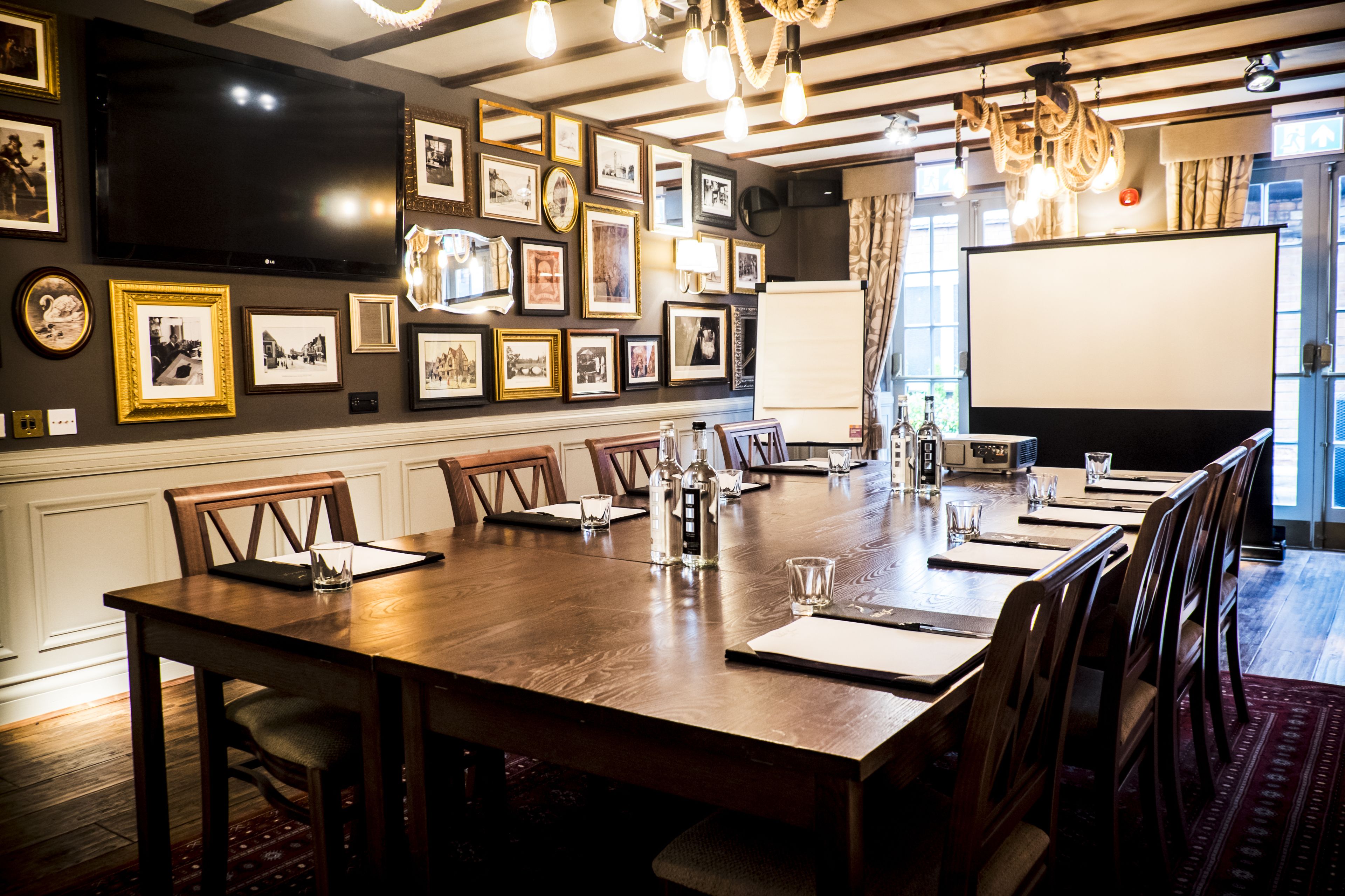 A conference room with a long table, chairs, a projector screen, and framed pictures on the walls.