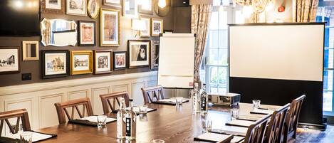 A conference room with a long table, chairs, framed pictures on the walls, and a projector screen.