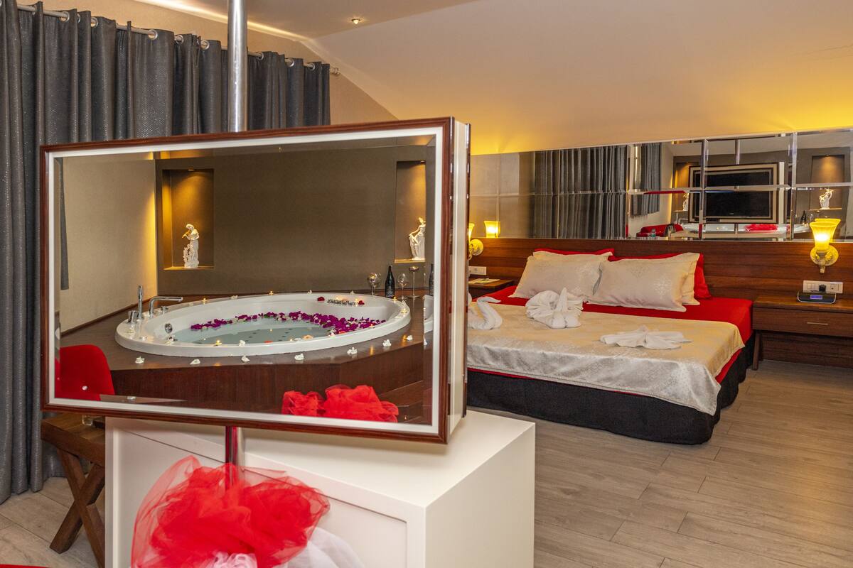 Habitación de hotel con un espejo grande, una cama con ropa de cama roja, una mesita de noche y un baño con bañera y espejo.