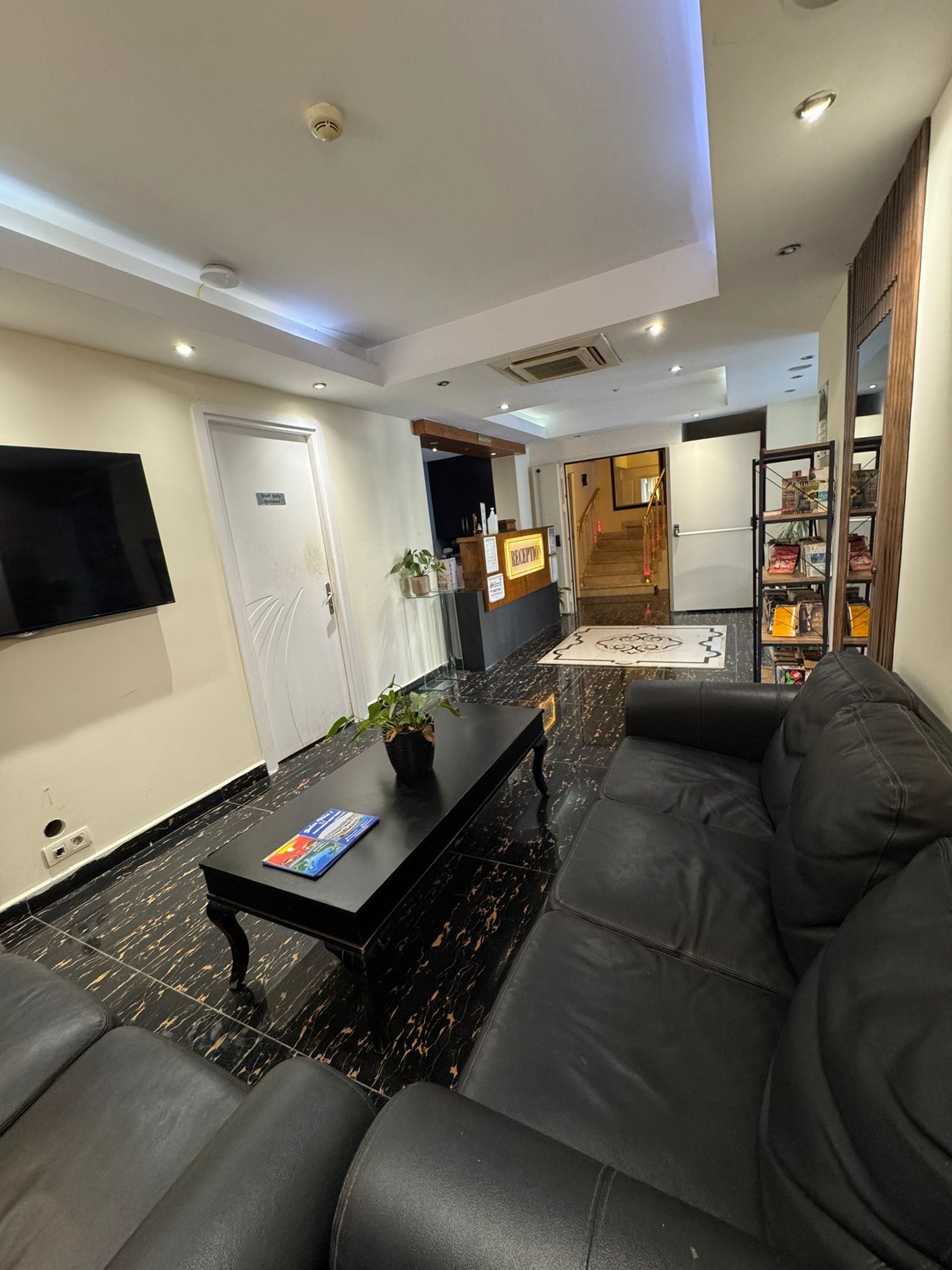 Una sala moderna con un sofá de cuero negro, un televisor de pantalla plana montado en la pared, un mostrador de recepción y una escalera que conduce a otro piso.