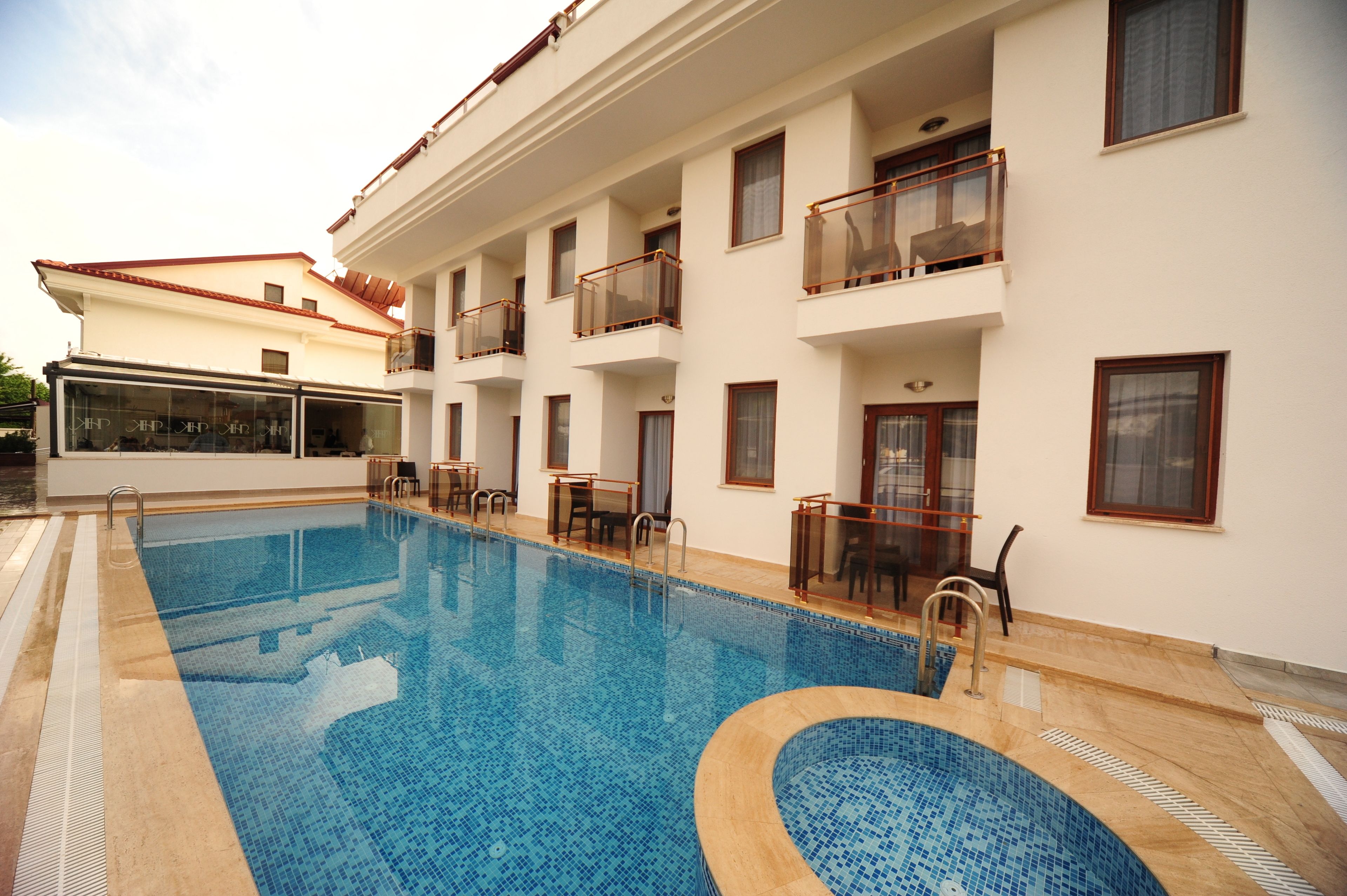 Una piscina con pavimento de baldosas, una zona cubierta con cristales y un edificio con balcones y ventanas.