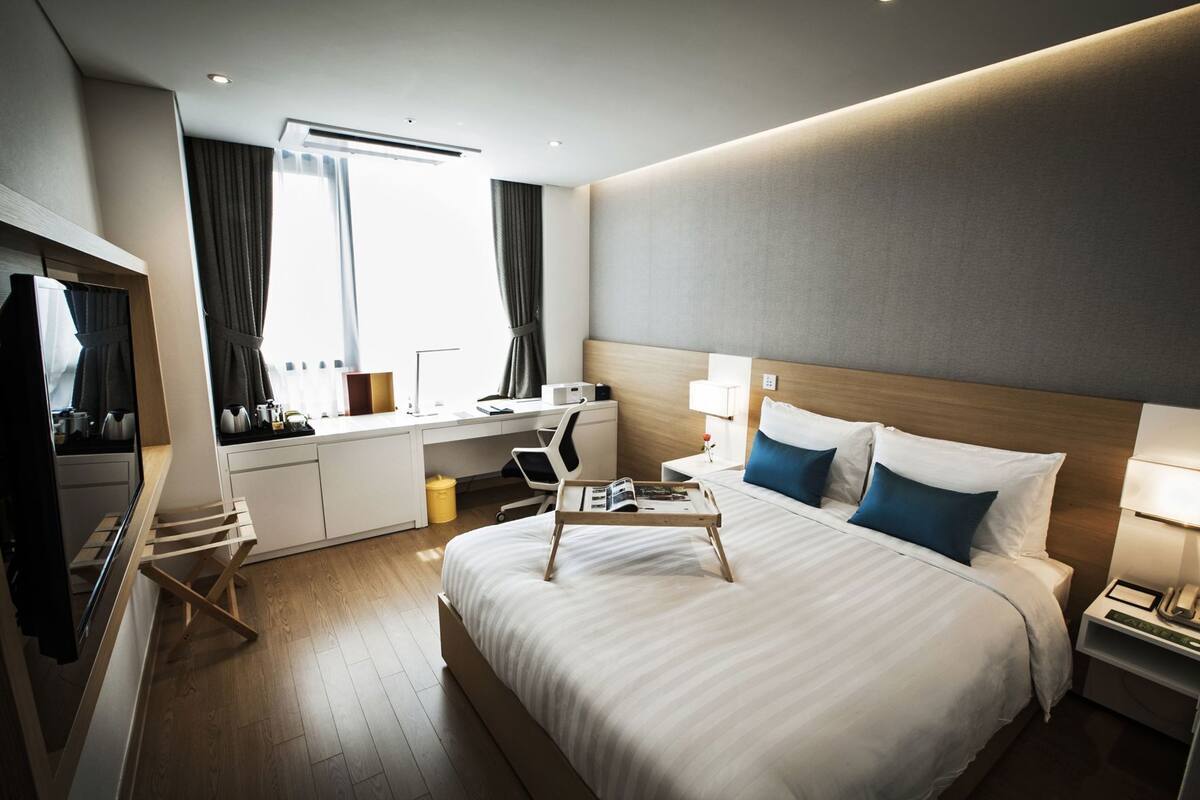 Una habitación de hotel con cama, escritorio, silla, televisor y ventana con cortinas.