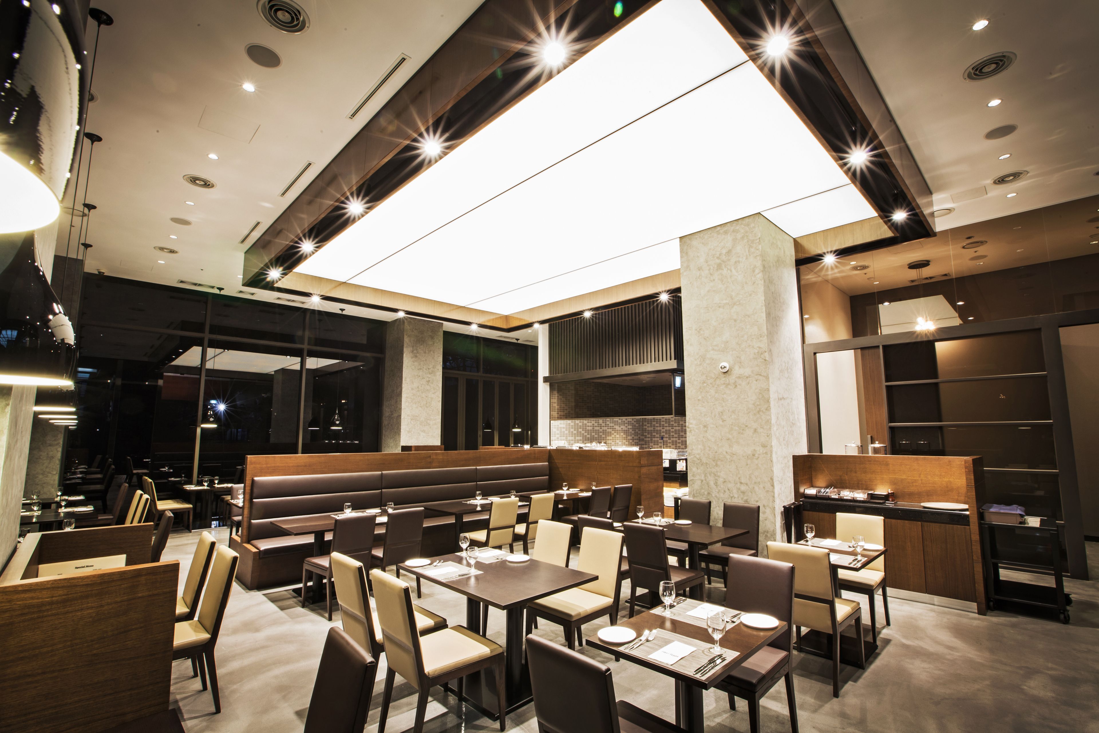 A modern restaurant with booths and tables, a large illuminated ceiling, and a kitchen area visible through a glass partition.