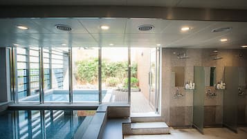 Una piscina interior moderna con paredes de vidrio, un área de spa con duchas y vistas a un jardín ajardinado.