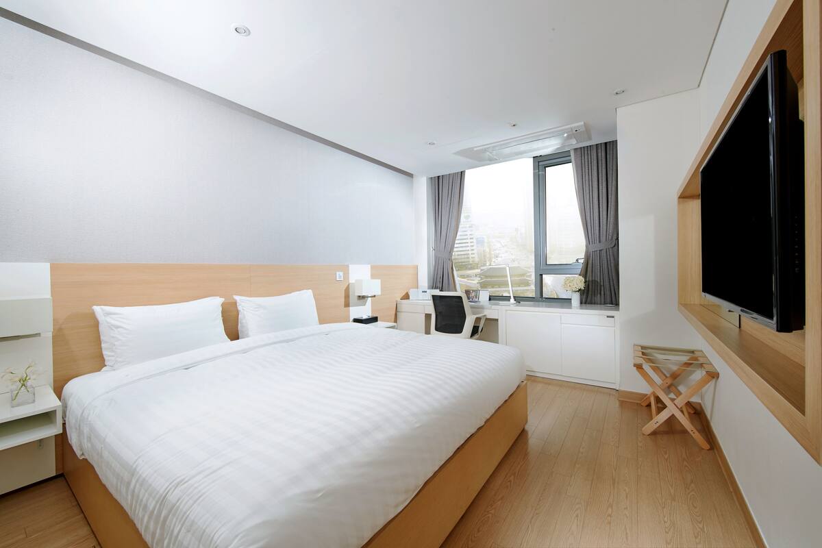 Una habitación de hotel moderna con una cama grande, un escritorio, una silla, un televisor y una ventana con cortinas.