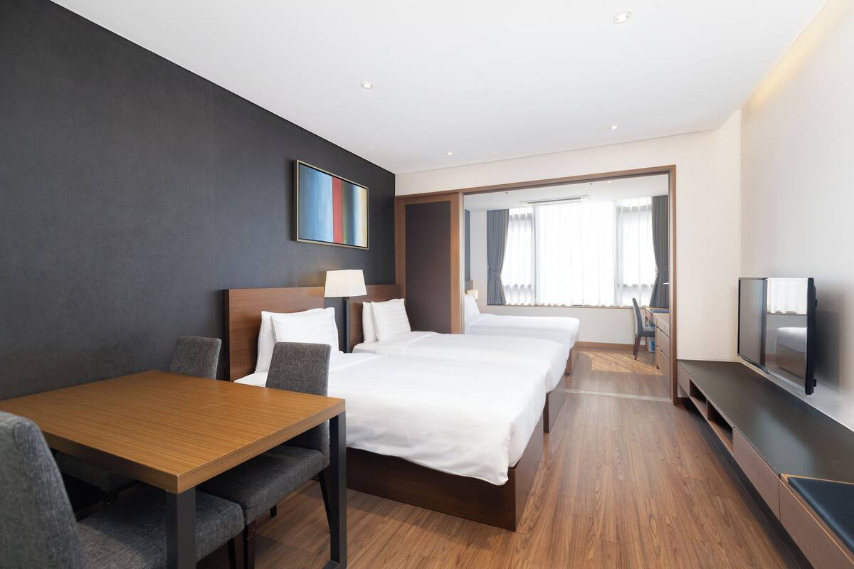 Una habitación de hotel con un escritorio de madera, dos camas, un televisor y un ventanal grande.