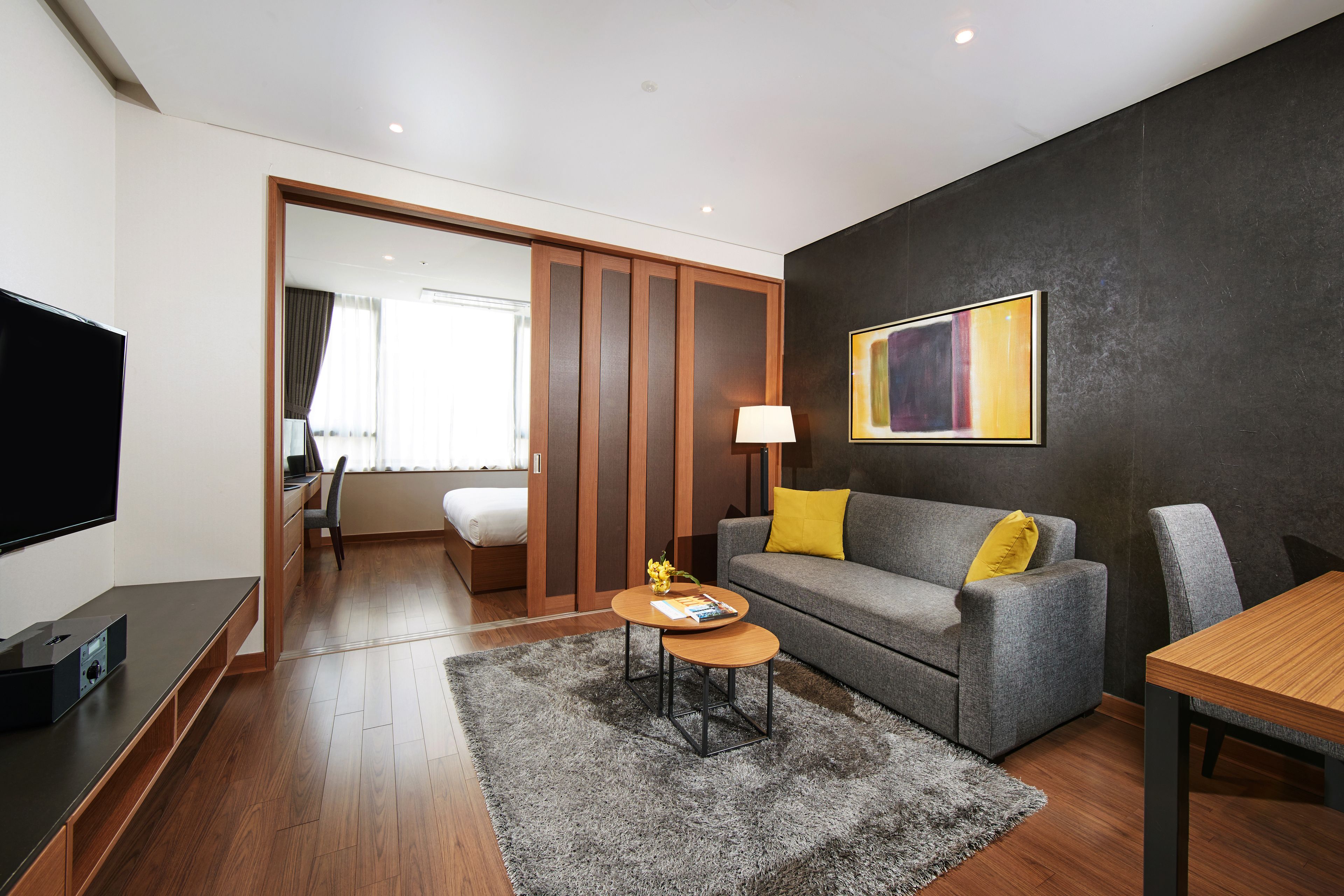 Ein modernes Hotelzimmer mit grauem Sofa, einem Holztisch, einem Fernseher und einem Gemälde an der Wand.